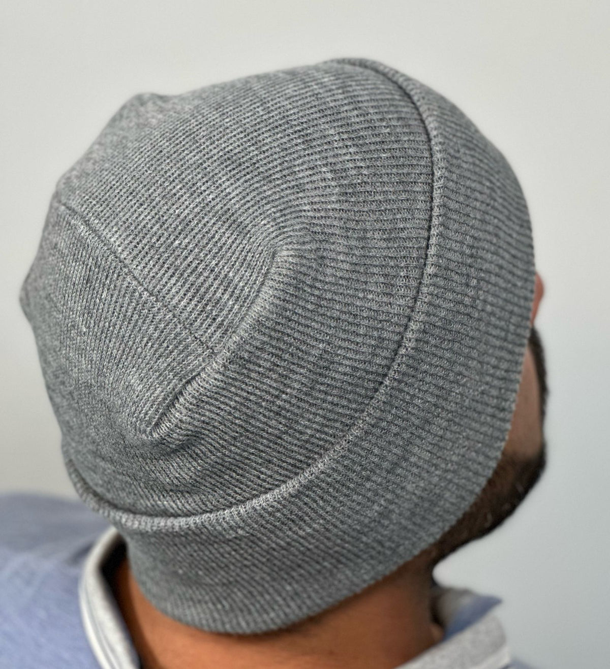 A gray hat featuring The Beanie Factory's lay-flat, irritation-free H-Stitch is modeled by a man with a black beard. The man is facing away from the camera so the H-Stitch is the focal point of the photograph.