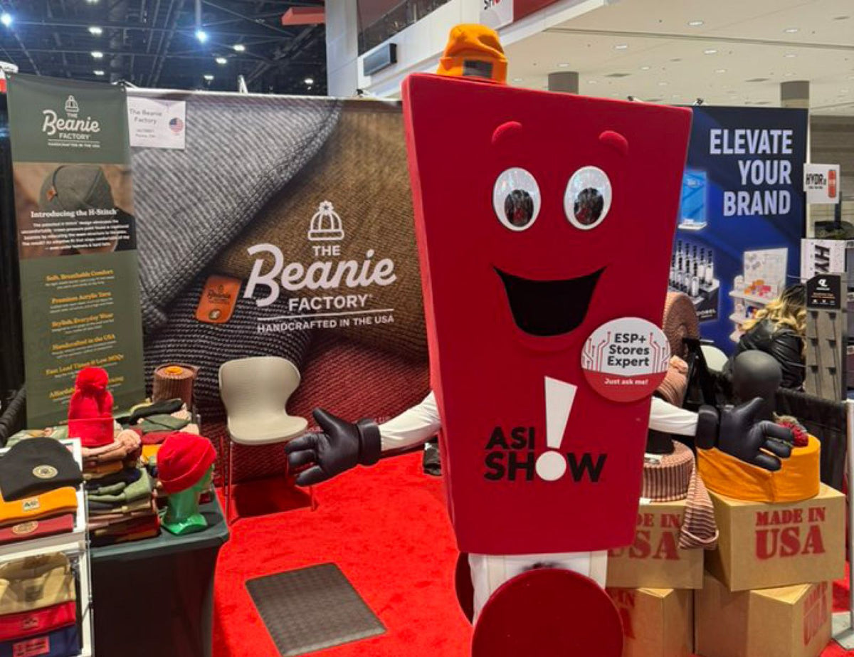 An image of The Beanie Factory booth at ASI trade show. The backdrop, pop-up banner, and multiple hat options are on display. The ASI Show mascot is standing at the booth. Multiple boxes say "Made In USA."