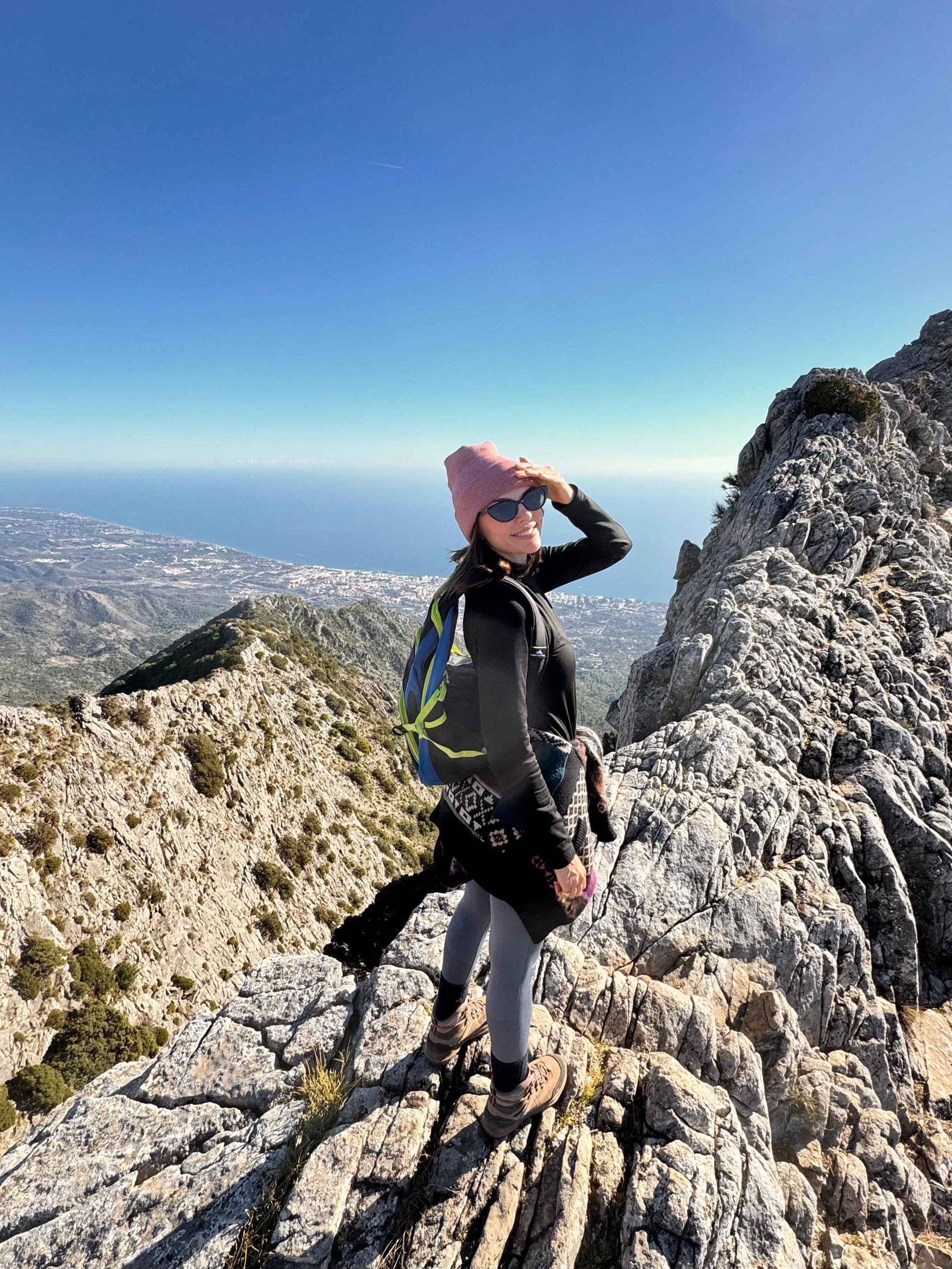 Person standing on a rocky mountain peak with a scenic view of the ocean and sky wearing a beanie made in USA. 